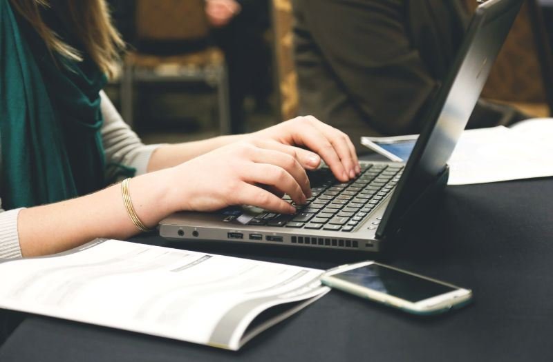 Mulher trabalhando em um texto para escrever no notebook, em uma mesa com papéis e um celular. 